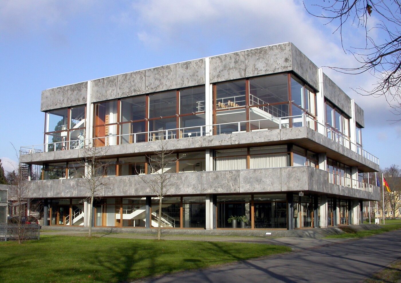 The Federal Constitutional Court building in Karlsruhe seen from outside on a winter day: a three-storey modernist block of pale concrete panels and full-height glazing, raised on slender columns, with bare trees and lawn in front and a German flag at the right.