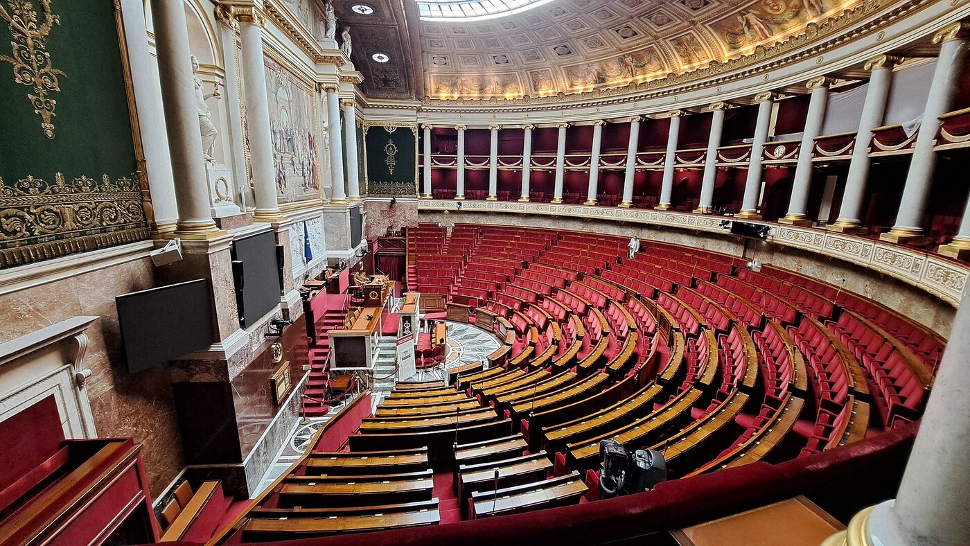Wide view of the semicircular debating chamber of the French National Assembly, tiers of red benches curving around the rostrum beneath a coffered ceiling.
