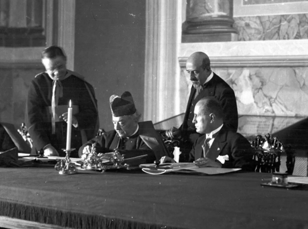 Black-and-white photograph of a cardinal in a biretta signing a document at a draped table, a man in a dark suit seated beside him and two others standing behind, in a marble-lined palace room