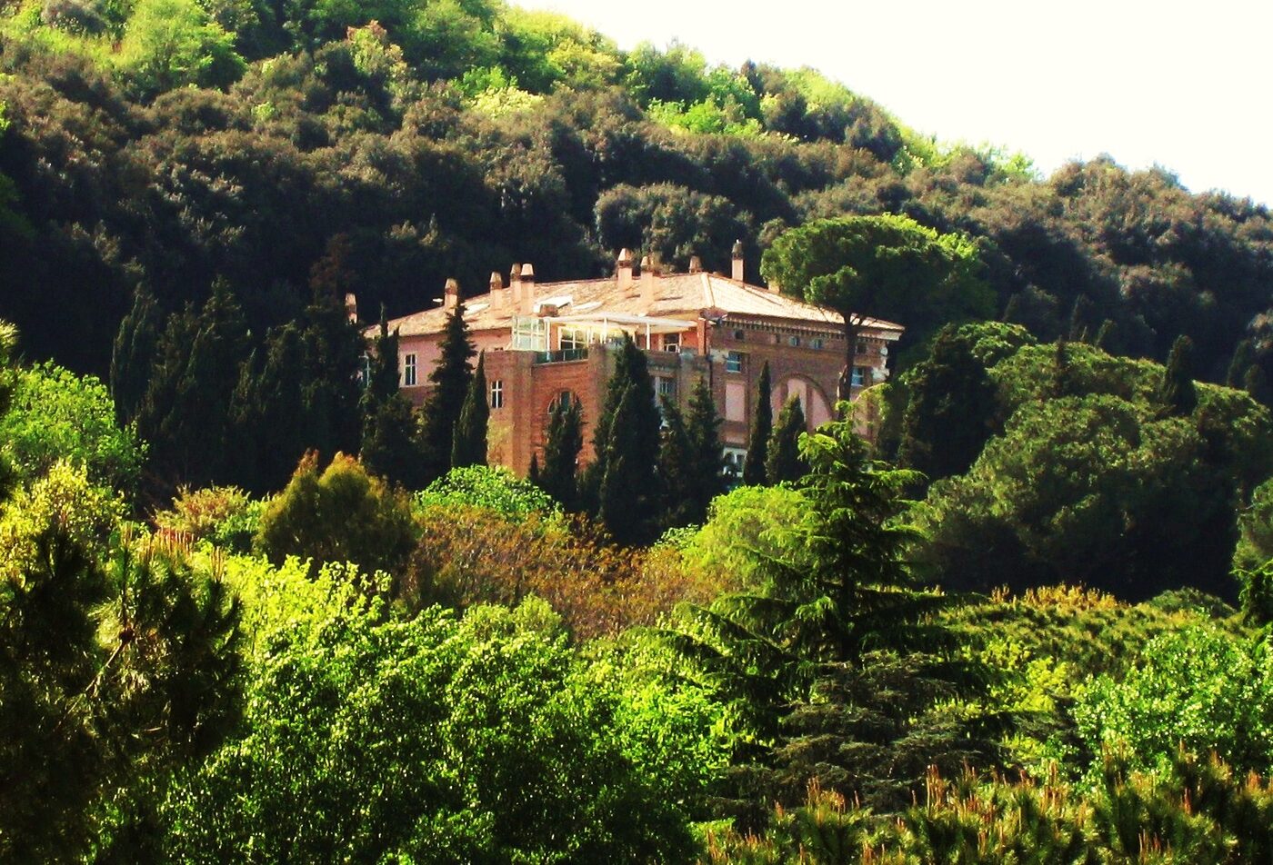 A Renaissance villa of pink render and brick, with a tall arched opening on its flank, standing among dense trees on a wooded hillside