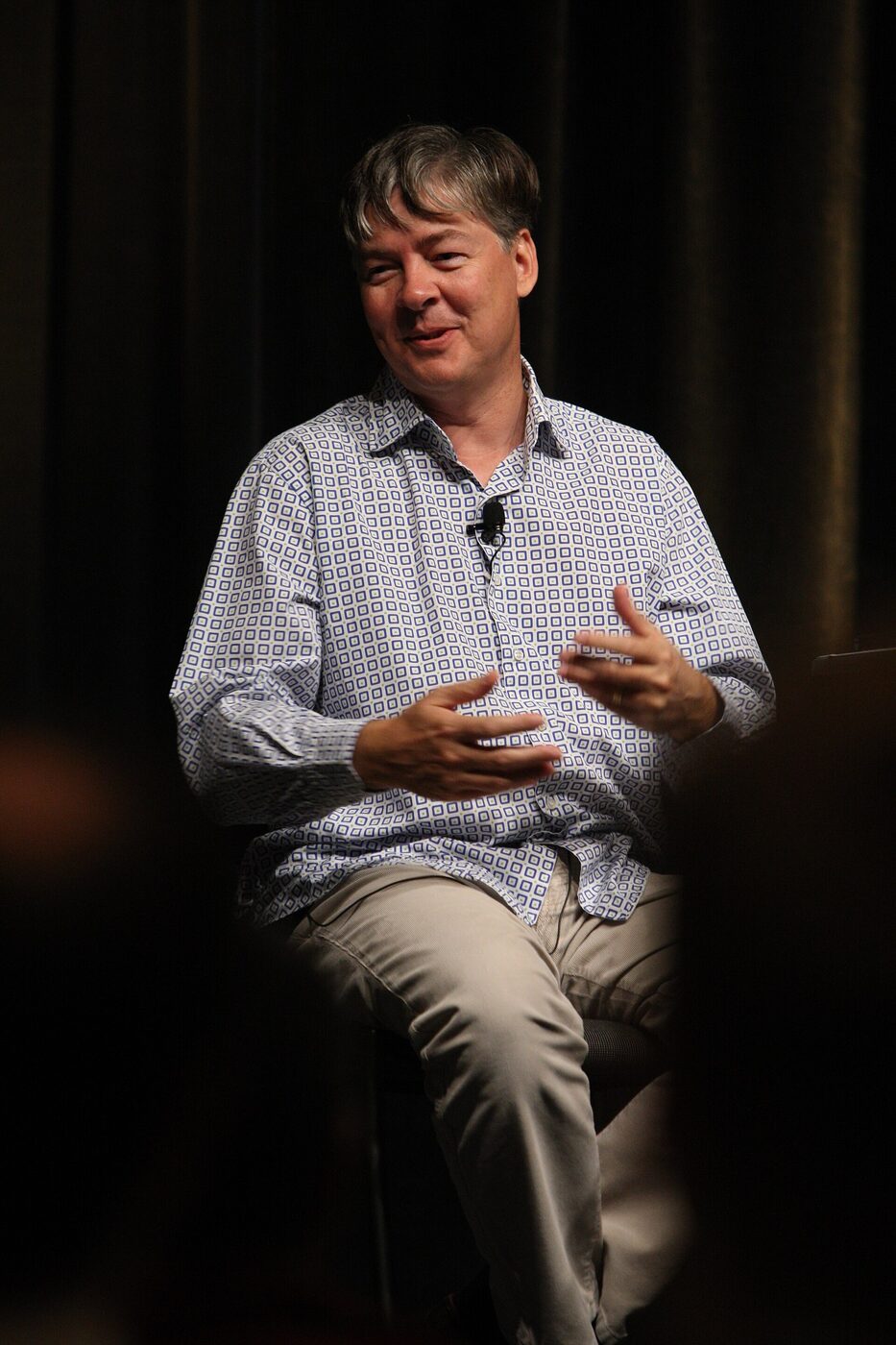 A man in a patterned shirt sits on a conference stage, speaking and gesturing with both hands against a dark backdrop.