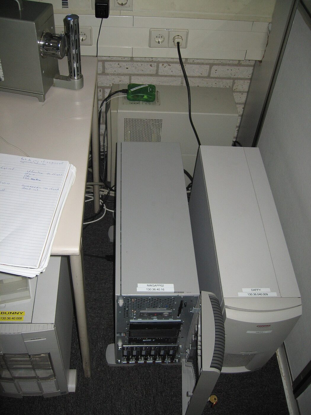 Two beige tower server computers standing on office carpet next to a desk, each with a paper label giving a host name and an IP address; one carries Compaq ProLiant branding.
