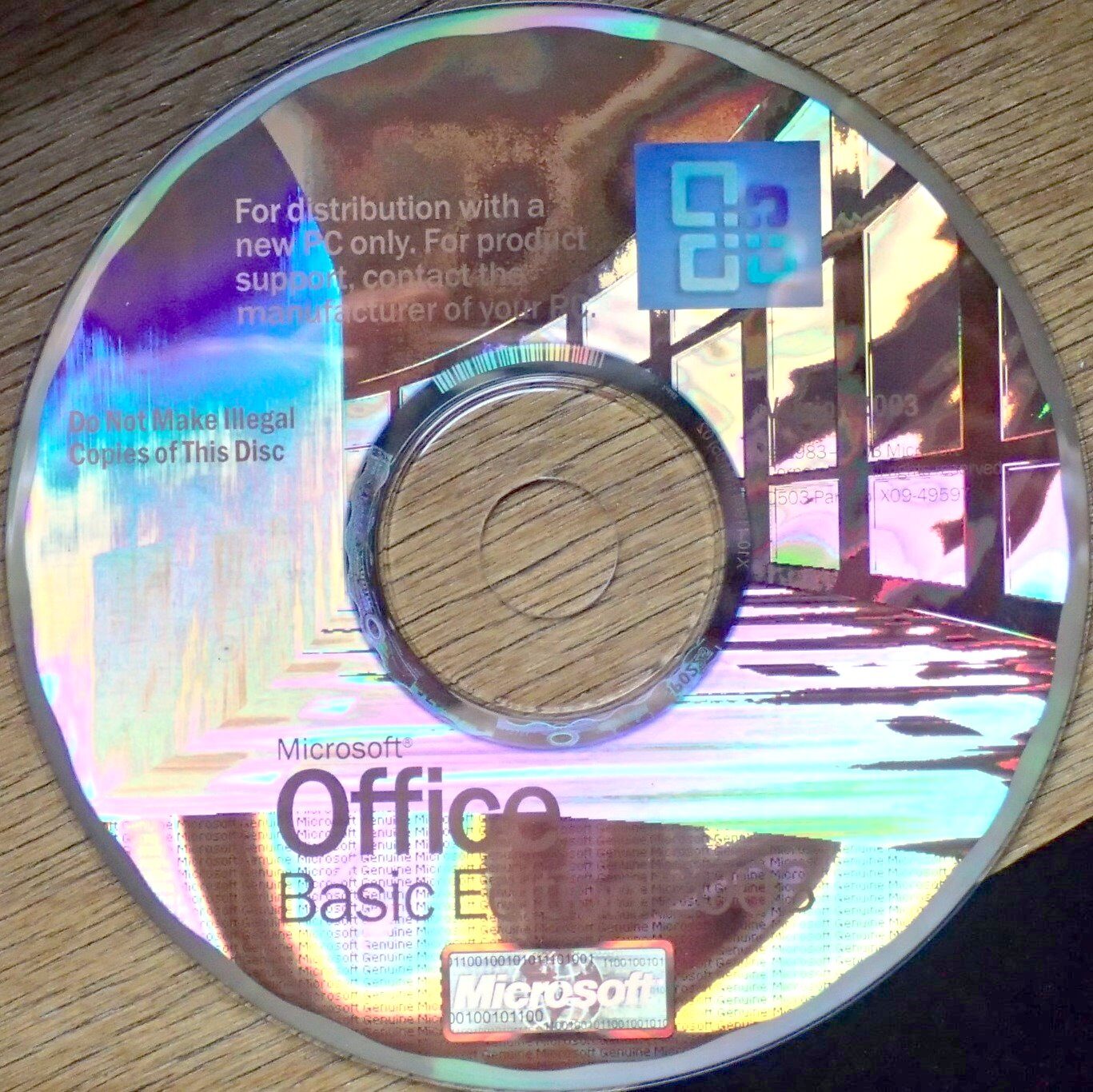 A Microsoft Office Basic Edition 2003 disc lying label-up on a wooden surface, printed with the Office logo and a notice reading 'For distribution with a new PC only'.