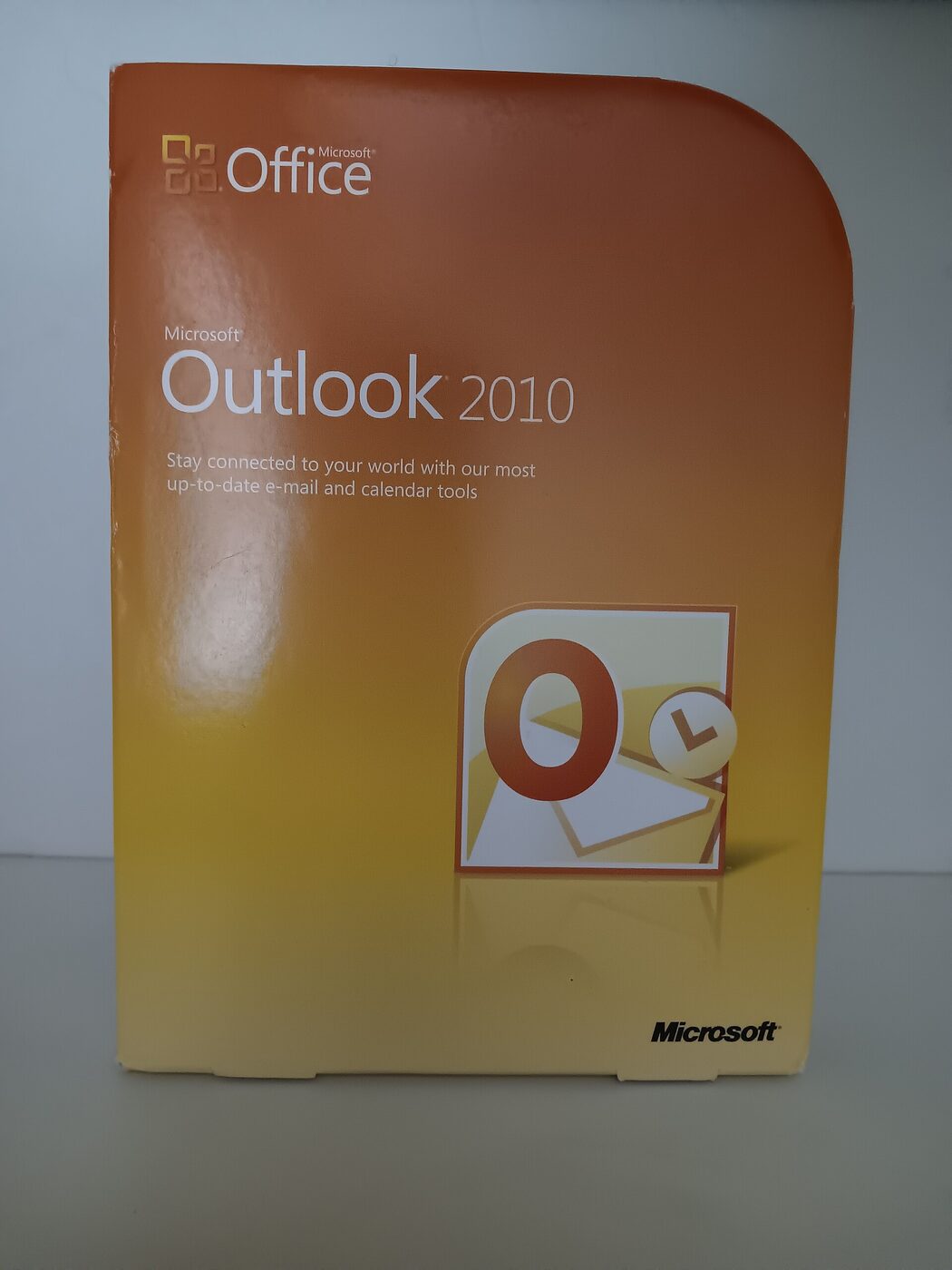 A retail software box for Microsoft Outlook 2010, orange and yellow, its front face showing the Office logo, the product name and the Outlook envelope-and-clock icon.