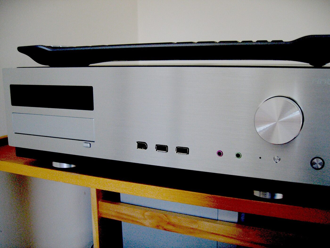 A slim silver hi-fi-style computer case standing on a wooden rack, with a large volume knob, three front USB sockets, pink and green audio jacks and a dark display panel; a black keyboard lies on top of it.