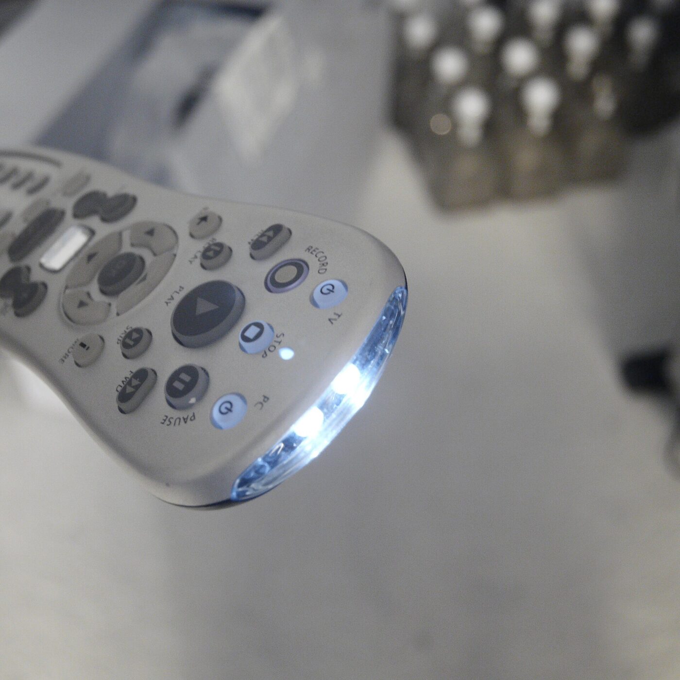 Close-up of the nose of a pale grey handheld remote control on a pale surface, its emitters glowing bright white, with buttons marked PLAY, PAUSE, STOP, RECORD and FWD and small round TV and PC power keys.