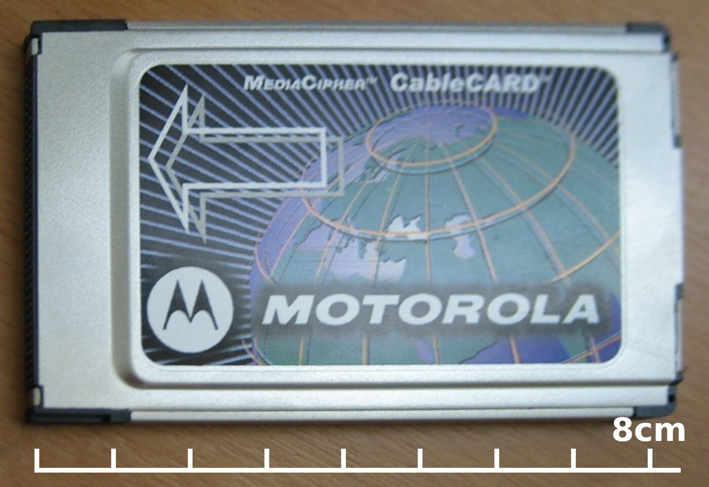 A silver metal-bodied PC Card lying face up on a wooden surface, its label printed with a globe, the words MediaCipher CableCARD and the Motorola name, with an eight-centimetre scale bar marked below it.