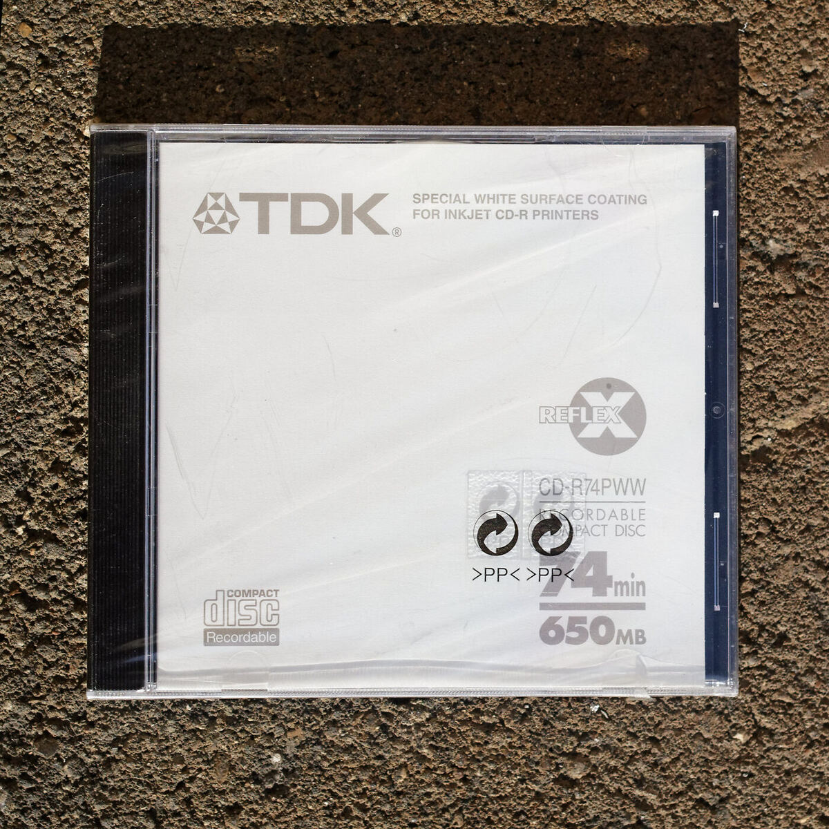 A single blank TDK CD-R in a shrink-wrapped jewel case, photographed from above on a concrete surface.