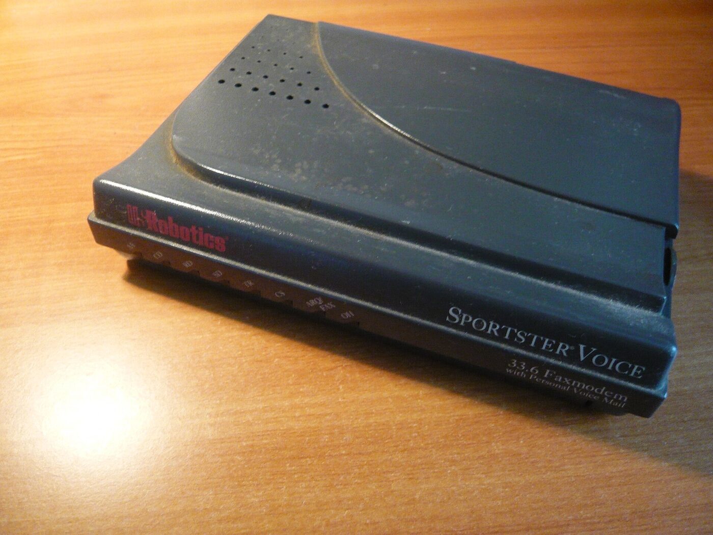 A black external dial-up faxmodem lying on a wooden desk, its front edge marked with a row of small labelled status lights and the words Sportster Voice 33.6 Faxmodem.