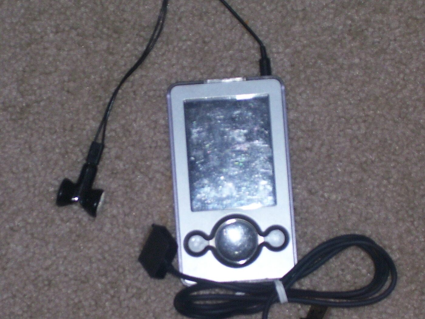A white rectangular portable media player with a screen above a circular control pad, lying on carpet beside a coiled black cable and a pair of earphones.
