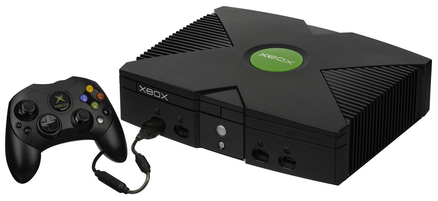 A black Microsoft Xbox console photographed from the front on a plain white background, with the smaller S-type controller resting in front of it.