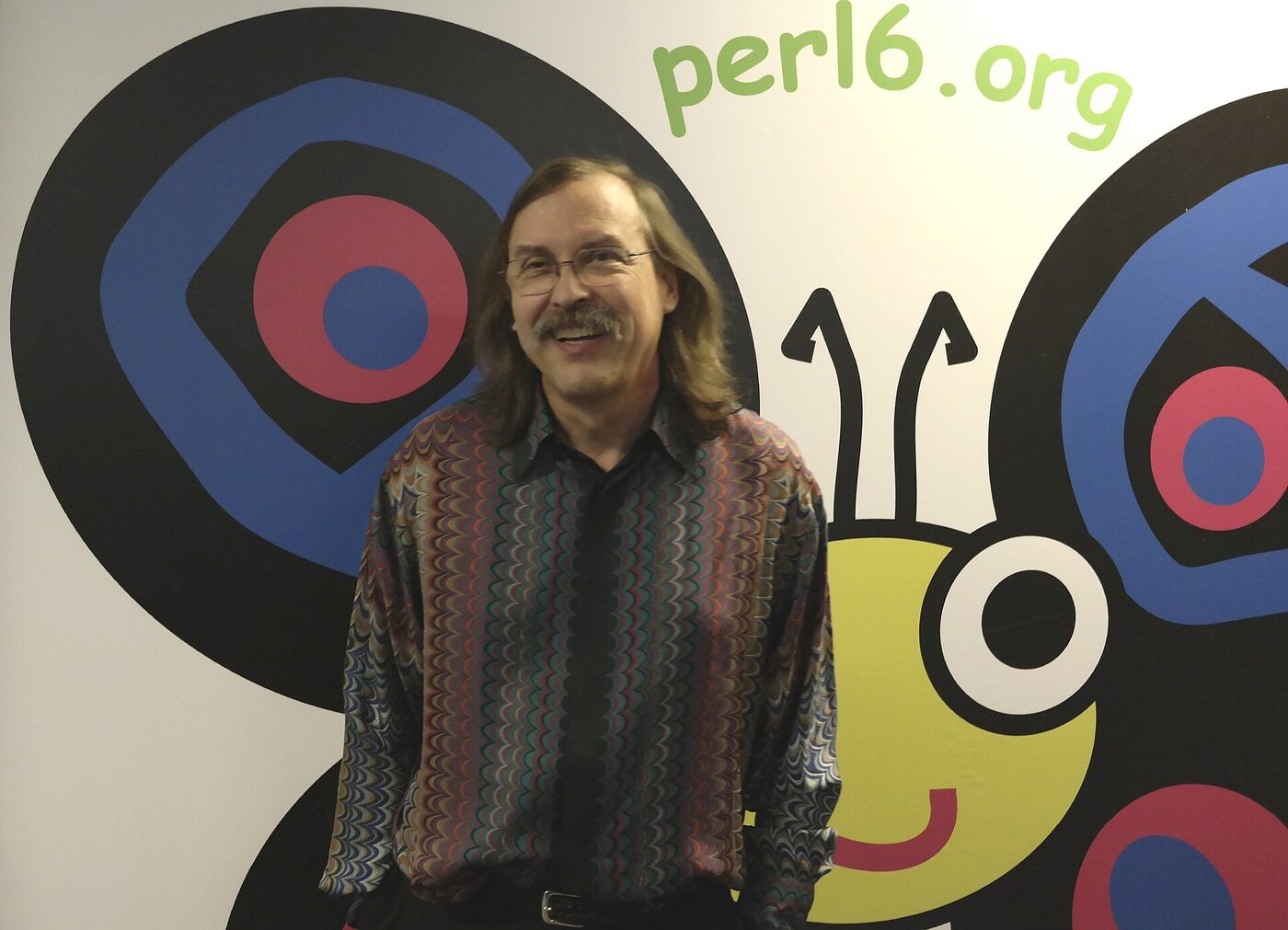 Larry Wall, a bespectacled man with long hair and a moustache in a patterned shirt, smiling in front of a wall painted with the Perl 6 butterfly logo and the text perl6.org.