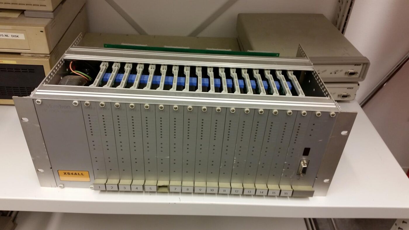 A grey rack-mount chassis on a depot shelf, its front filled with sixteen numbered modem cards with rows of indicator holes, an orange XS4ALL sticker at the lower left and older equipment stacked behind.