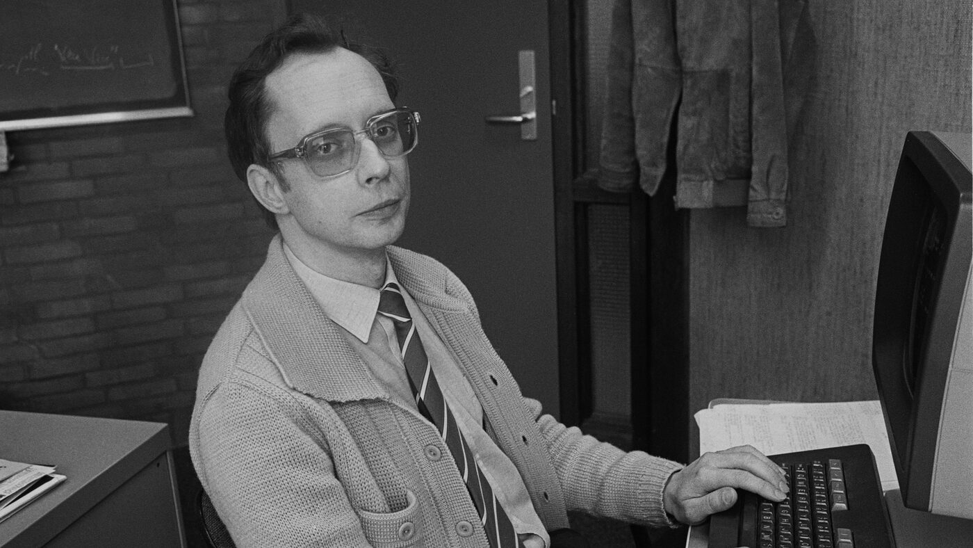Black-and-white photograph of a bespectacled man in a cardigan and tie seated at an office desk, one hand on a terminal keyboard beside a monitor and a stack of printout.
