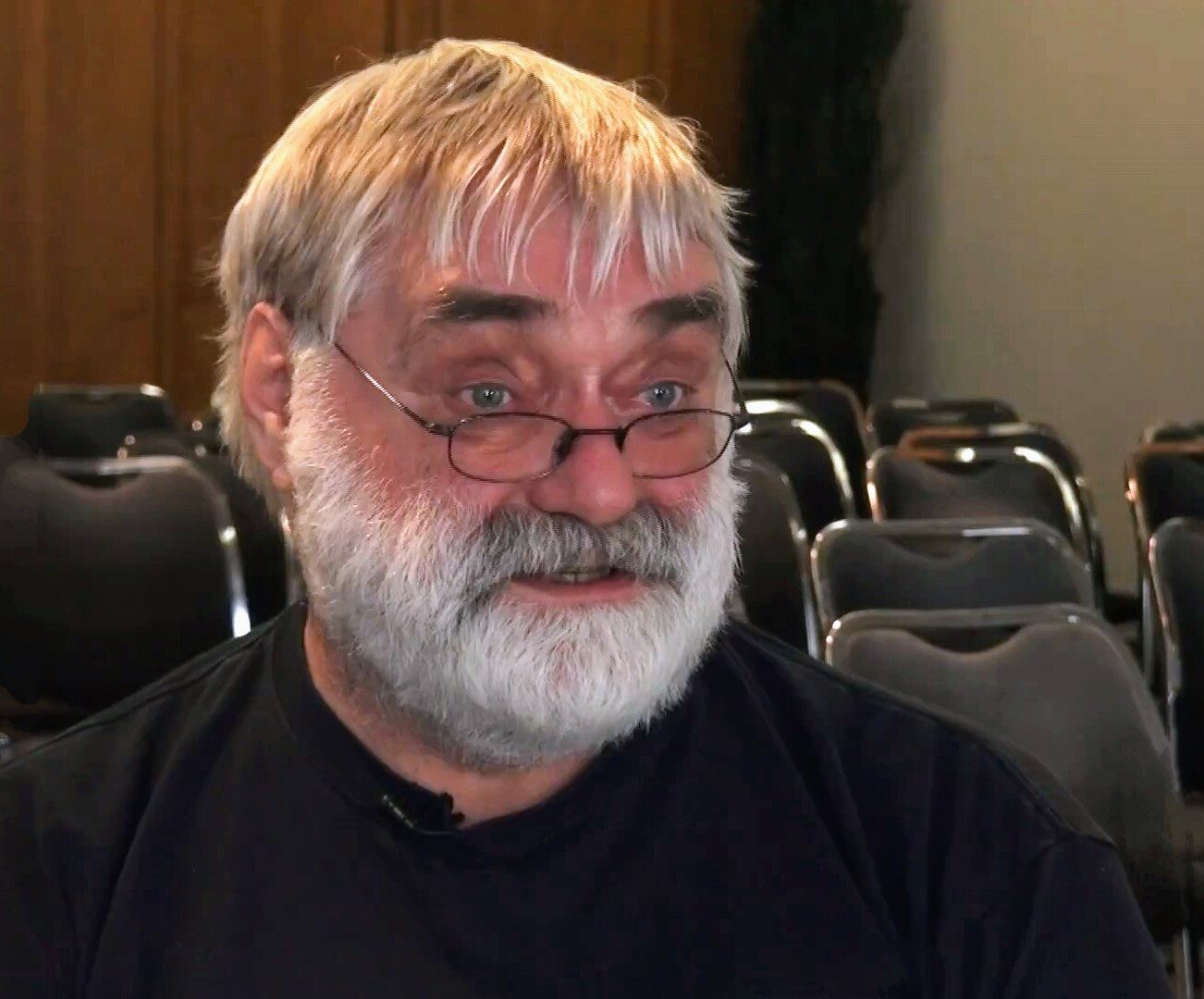 Still from a video interview: a white-bearded man in half-moon glasses and a dark T-shirt, sitting in front of rows of empty conference chairs.