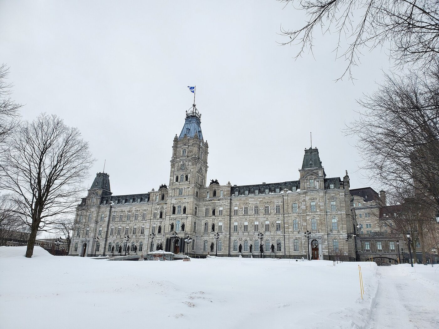 A large nineteenth-century Second Empire stone parliament building with a tall central clock tower flying the Quebec flag, seen from the front across deep snow, with bare trees on either side under an overcast sky.
