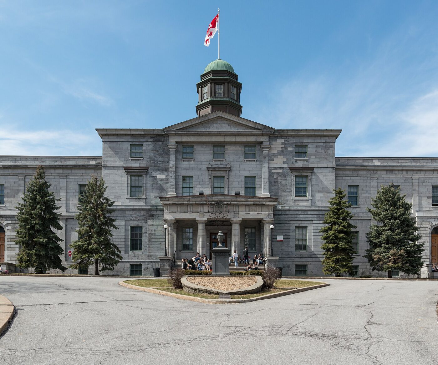 A grey stone neoclassical university building with a central portico and a green-domed octagonal cupola flying a flag, seen across a paved forecourt under a blue sky, with students sitting on the entrance steps.