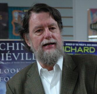 A man with a full grey beard, in a herringbone tweed jacket and open-necked white shirt, speaking, photographed in a bookshop with promotional posters for other novels on the wall behind him.