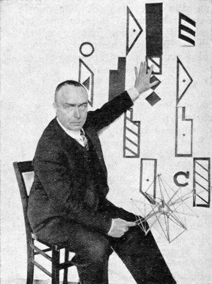 Black and white photograph of a seated man in a dark suit reaching up towards a wall display of black geometric notation symbols, holding a wire polyhedral model in his other hand.