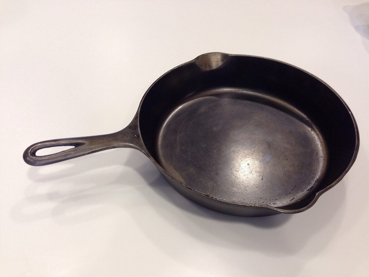 A well-used black cast-iron frying pan with a long looped handle and pouring lips, photographed at an angle on a pale surface.