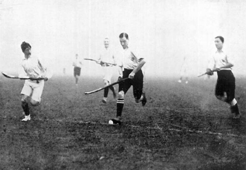 Black-and-white photograph of five hockey players in white shirts and knee-length shorts running on a bare grass field, each carrying a curved stick, the ball on the ground at the feet of the nearest player.