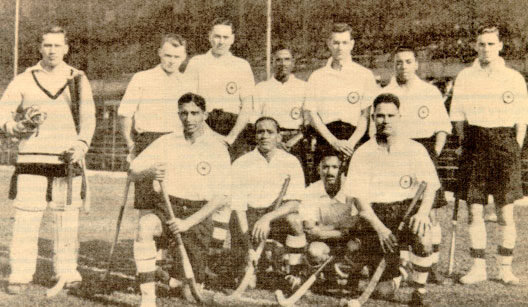 Sepia group photograph of the 1928 Indian Olympic hockey squad posed in rows in light shirts and dark shorts, several of the players holding sticks.