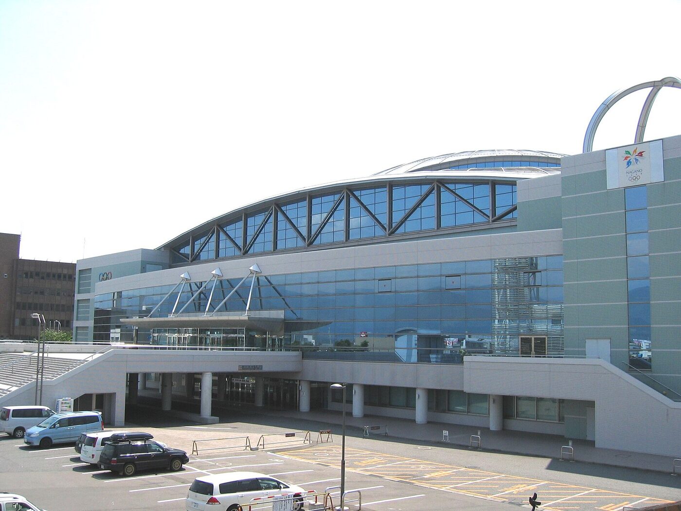 Exterior of a large arena with a curved glass-and-steel frontage and an arched roof, seen across a car park on a bright day, with the Nagano 1998 Olympic emblem mounted high on the right-hand wall.