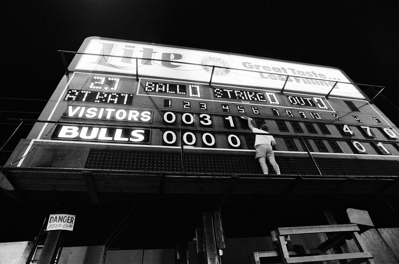 Night photograph looking up at a floodlit manual baseball scoreboard reading VISITORS and BULLS, with a man standing on the catwalk reaching up to change an inning number.