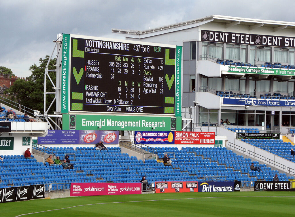 A large open cricket scoreboard showing the total, wickets, overs and individual batsmen's scores, with a stand and floodlight pylon behind it.