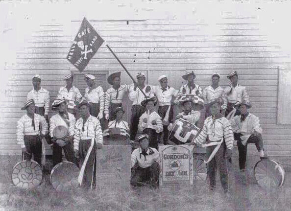 A black and white photograph of a steelband posed in two rows outside a wooden building, players in striped sleeves and peaked caps standing behind tuned metal drums, with a banner reading Hell's Gate Steel Band.