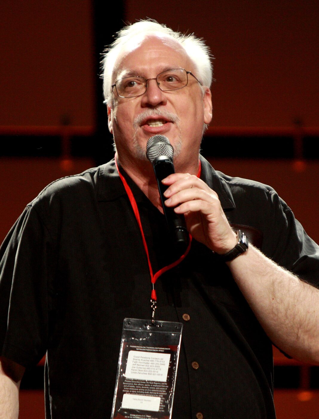 A bearded man in a dark shirt speaking into a microphone at a convention panel.