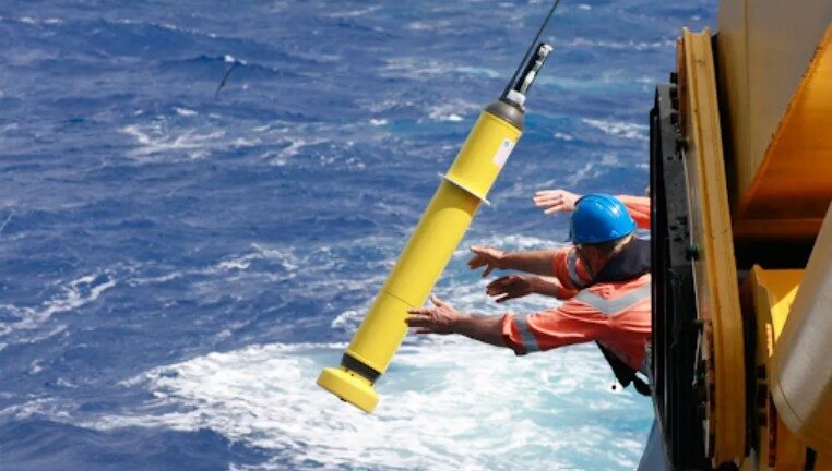 A yellow cylindrical instrument with a thin antenna falling towards the open sea beside a ship's hull, two crew members in orange overalls, one in a blue hard hat, reaching out after it.