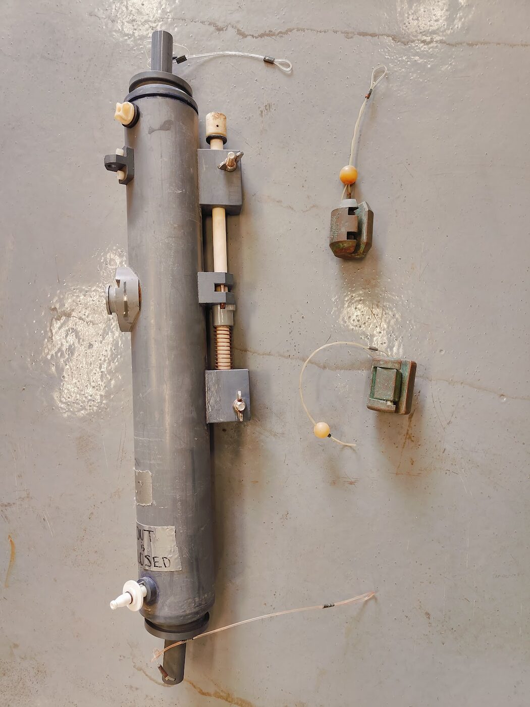 A long cylindrical plastic water-sampling bottle standing with rubber end caps, a spring-loaded release mechanism and lanyards, with small cylindrical metal messenger weights beside it.