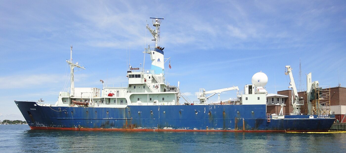A blue-hulled research ship with a white superstructure, a radome and cranes on the after deck, lying alongside a dock.