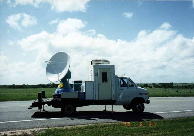 A flatbed truck carrying a large white parabolic radar dish on a rotating mount, parked on a roadside verge beside open grassland under a partly cloudy sky, with a camera date stamp in the corner.