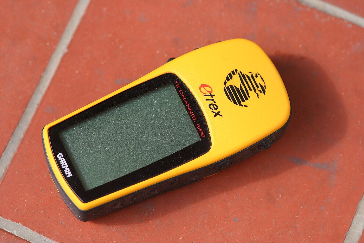 A yellow handheld GPS receiver lying face up on red floor tiles, its monochrome screen blank, with Garmin, eTrex and “12 channel GPS” markings visible on the case.