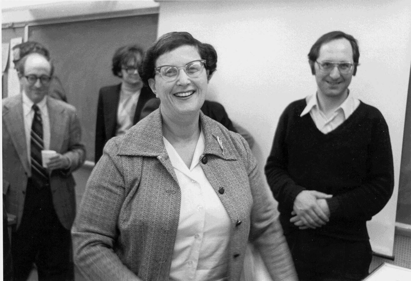 A photograph of a woman standing and speaking in a lecture room, holding papers.