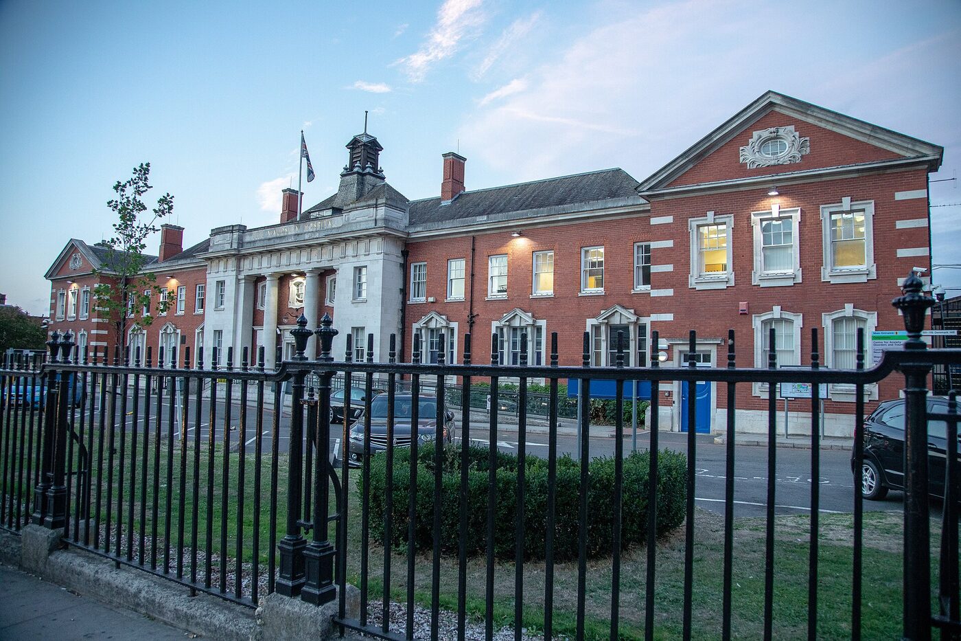 A long red-brick hospital building with white stone dressings and a columned entrance portico, seen across a road behind iron railings at dusk.