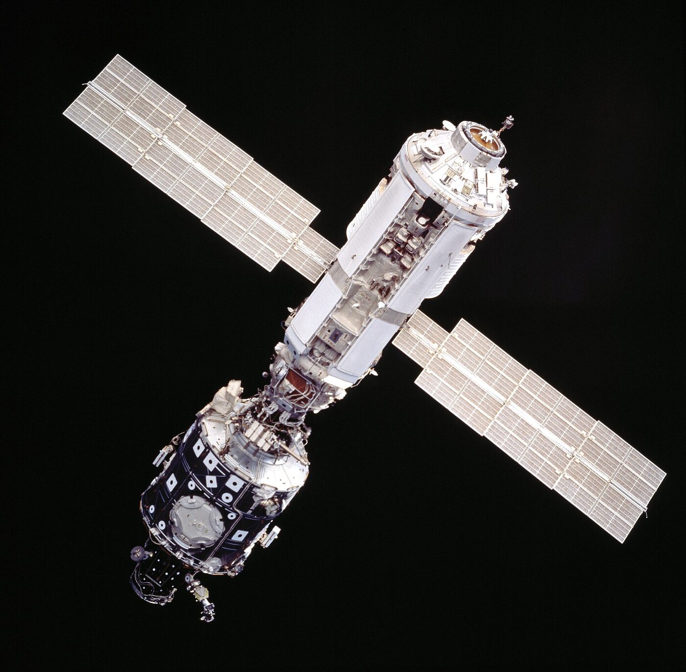Two connected cylindrical space station modules with a pair of deployed solar arrays, silhouetted against black space.