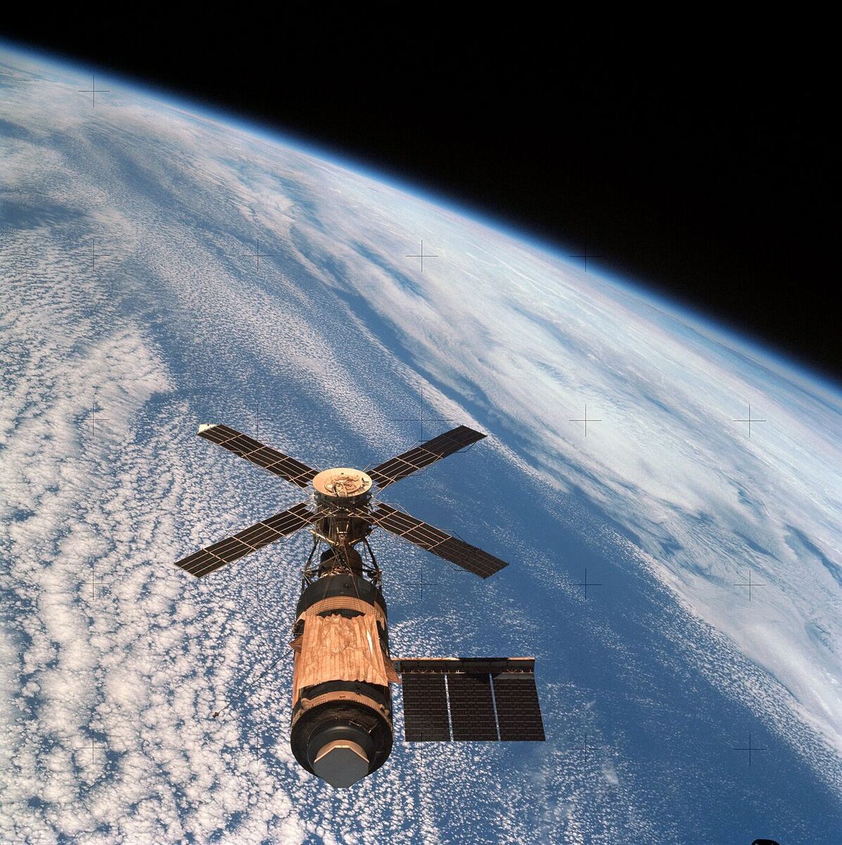 A space station made from a converted rocket stage, with one large solar array on one side, a windmill of four smaller arrays at the front and a gold-coloured sheet covering the main cylinder, seen against black space with the curve of the Earth behind.