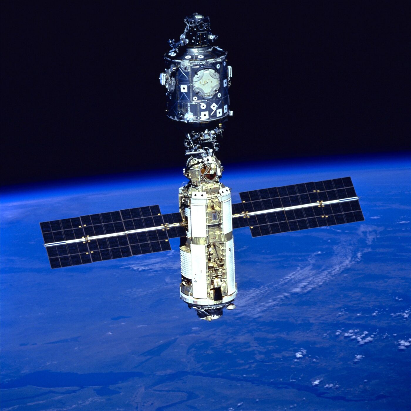 Two docked space station modules in orbit, one with a pair of long solar arrays extended either side, seen against the blue curve of the Earth below and black space above.