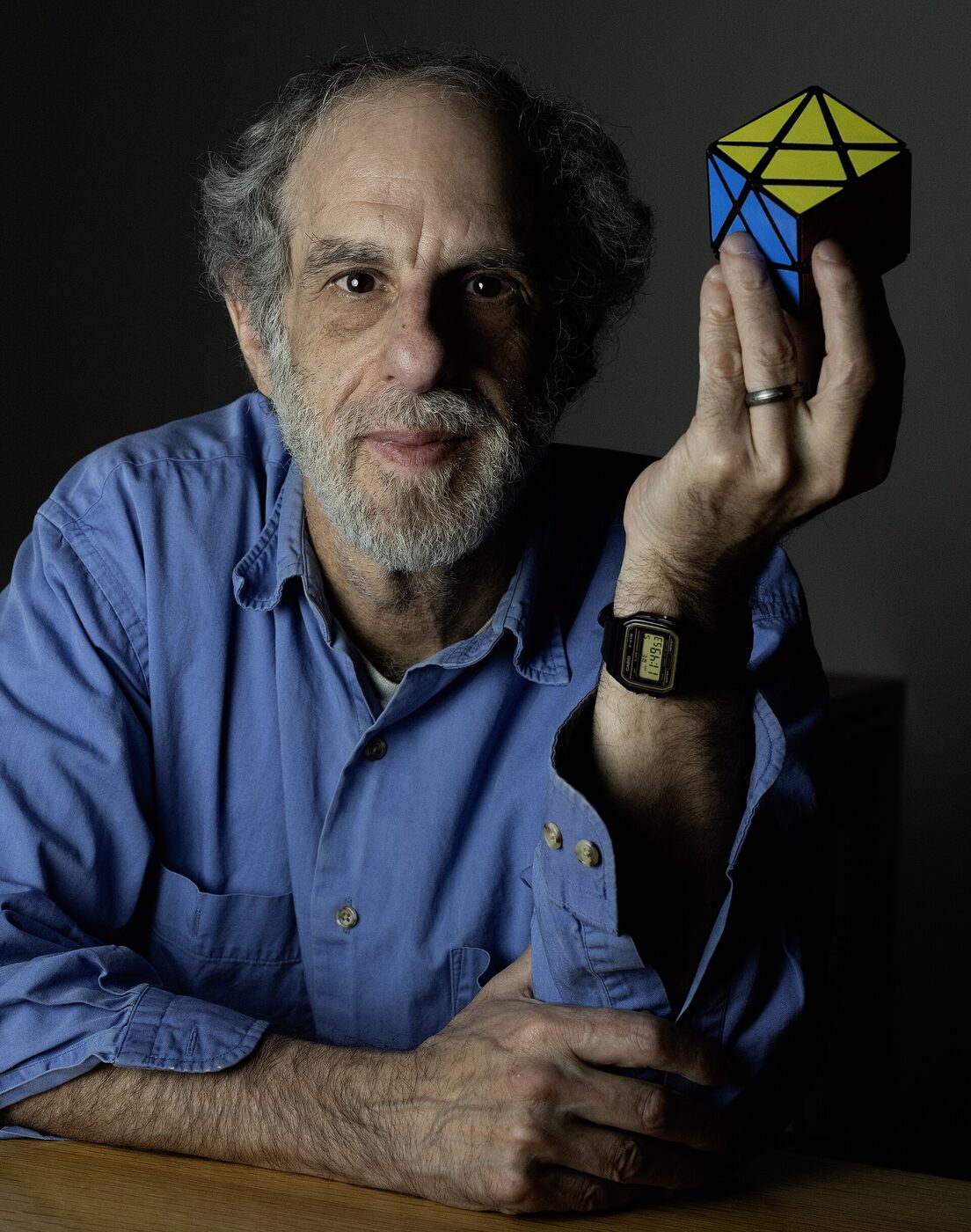 A bearded, grey-haired man in a blue shirt seated at a table, holding up a small coloured twisty puzzle in one hand.