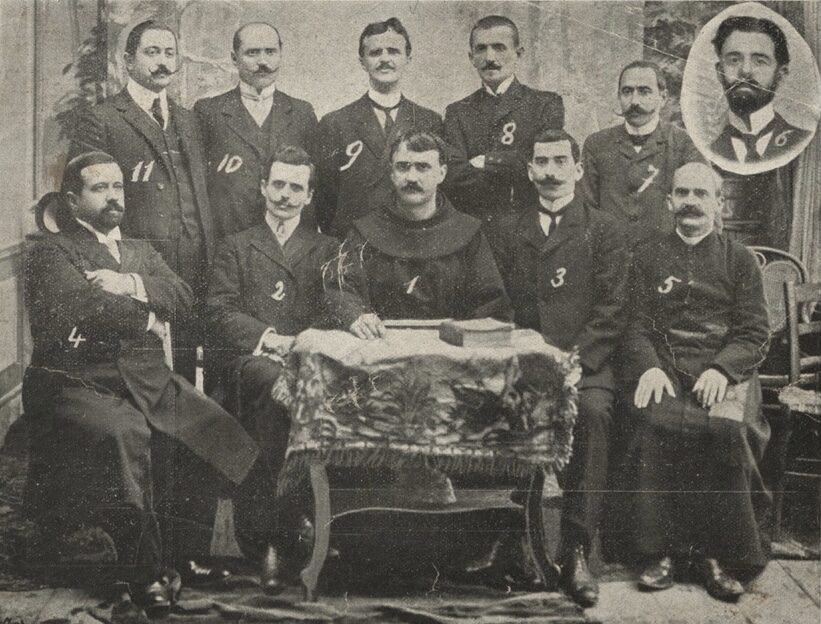 A formal group photograph from 1908 of about a dozen men in early twentieth-century dress, some in clerical or traditional costume, arranged in seated and standing rows.