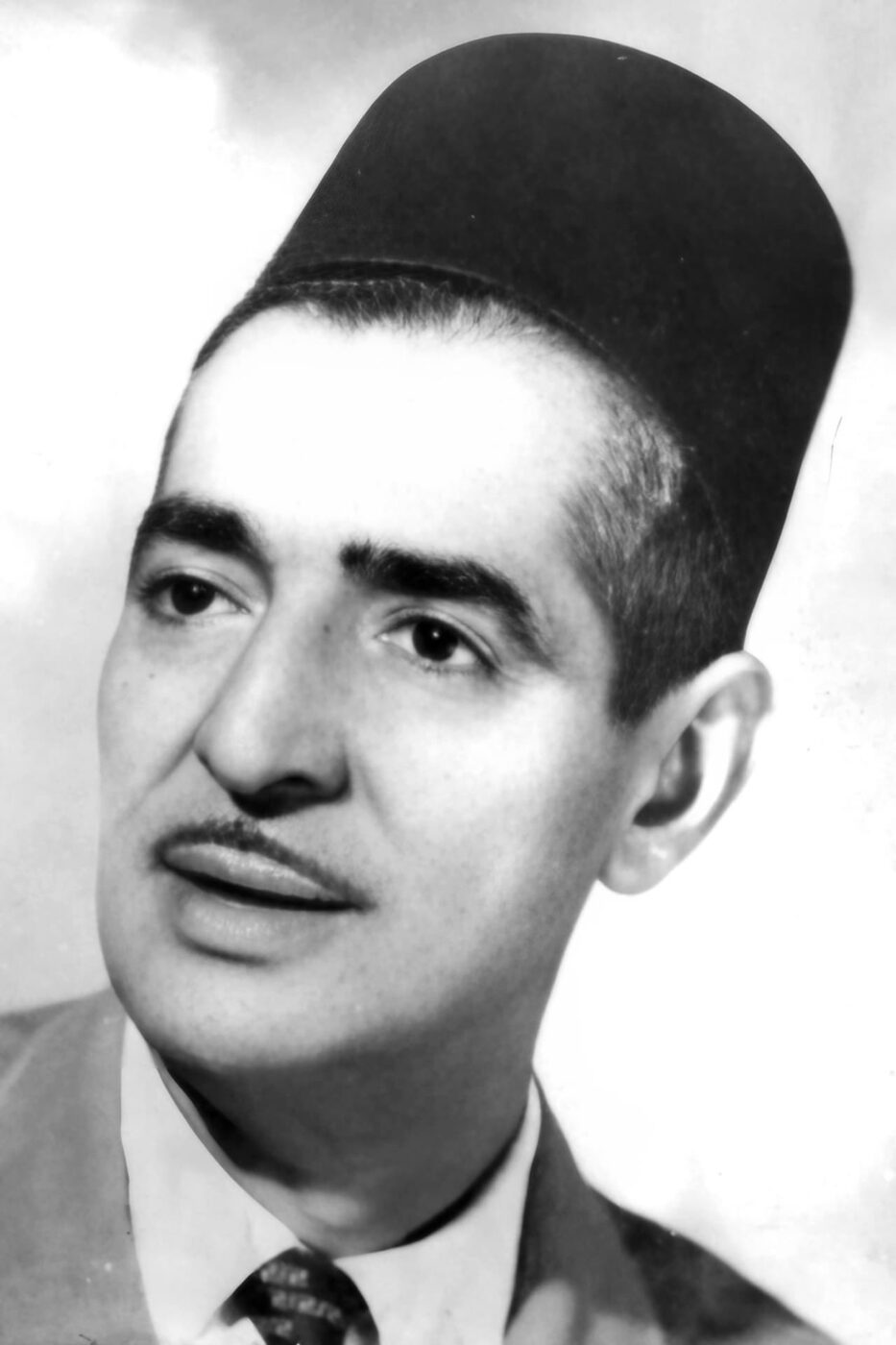 Black-and-white studio portrait, head and shoulders, of a moustached man in a suit and patterned tie wearing a tall brimless felt hat.