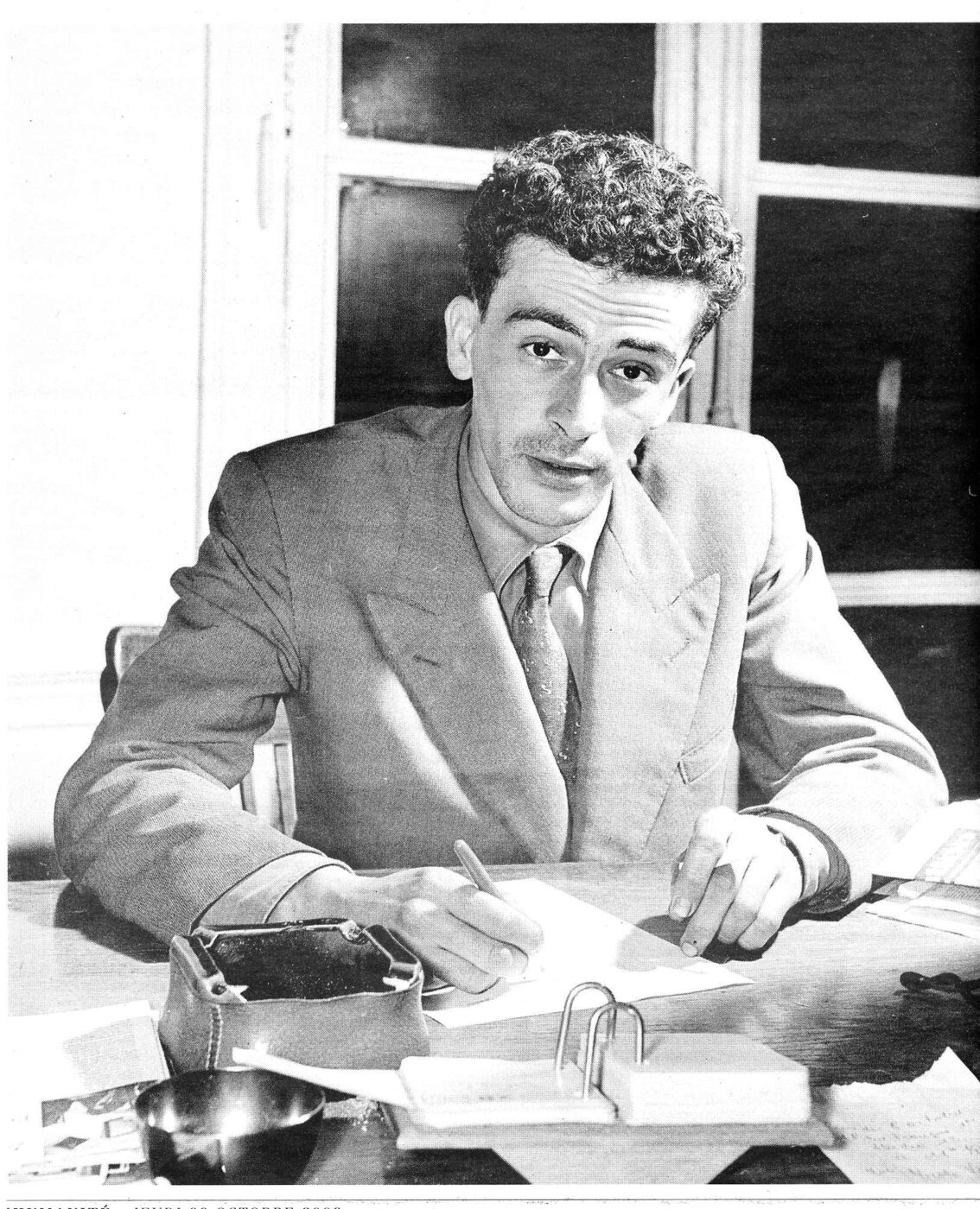 Black-and-white photograph of a young man in a loose suit seated at a desk, pen in hand over a sheet of paper, with an ashtray, papers and a stapler in front of him.