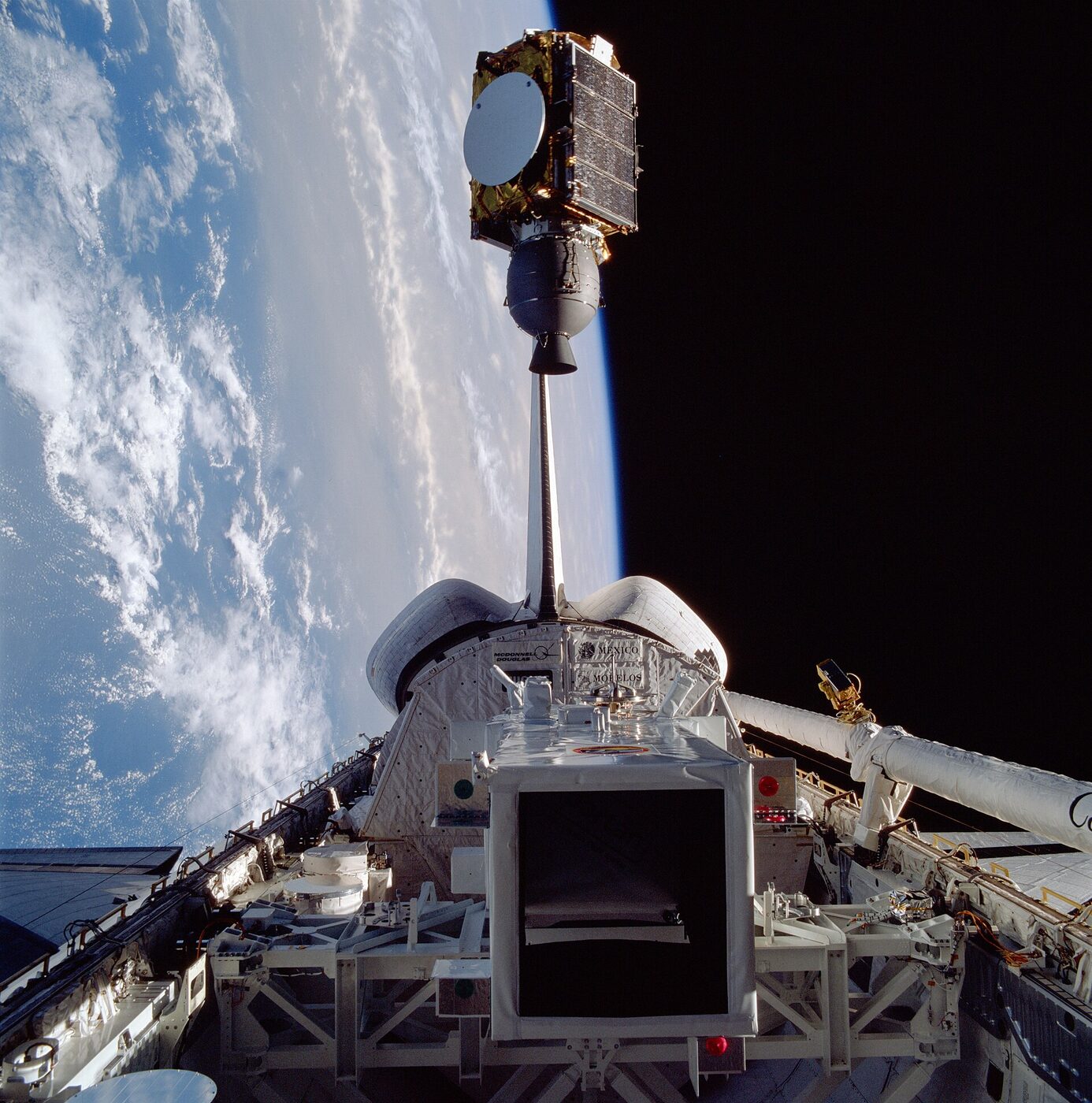 A drum-shaped communications satellite drifting away from a space shuttle's open payload bay, with the cloudy surface of the Earth at left.
