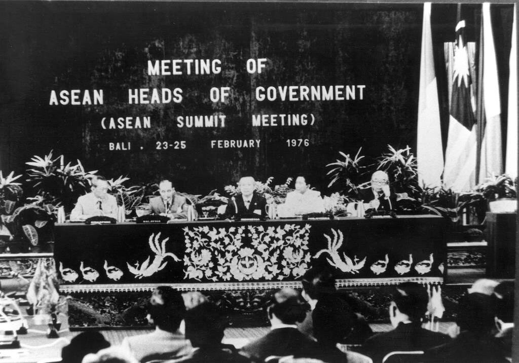 Black-and-white photograph of five heads of government seated at a carved dais beneath a banner reading MEETING OF ASEAN HEADS OF GOVERNMENT (ASEAN SUMMIT MEETING), BALI, 23-25 FEBRUARY 1976, with flags at the right and an audience in the foreground.