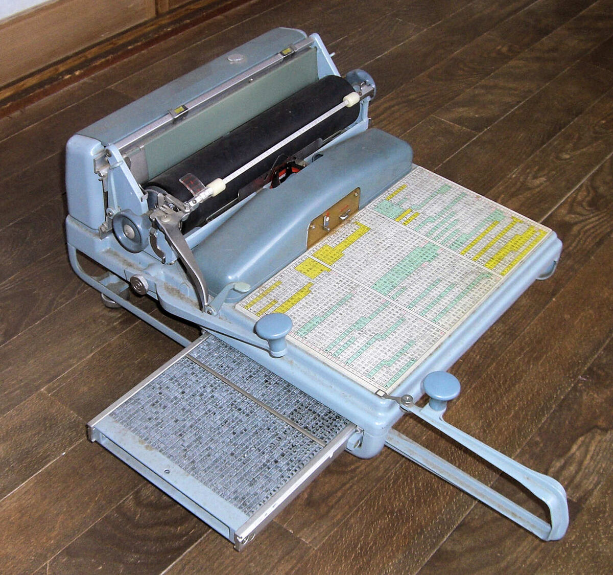 A desk-sized mechanical Japanese typewriter with a wide drawer holding several thousand small metal type pieces, a colour-coded printed character index beside it and a paper carriage at the rear.