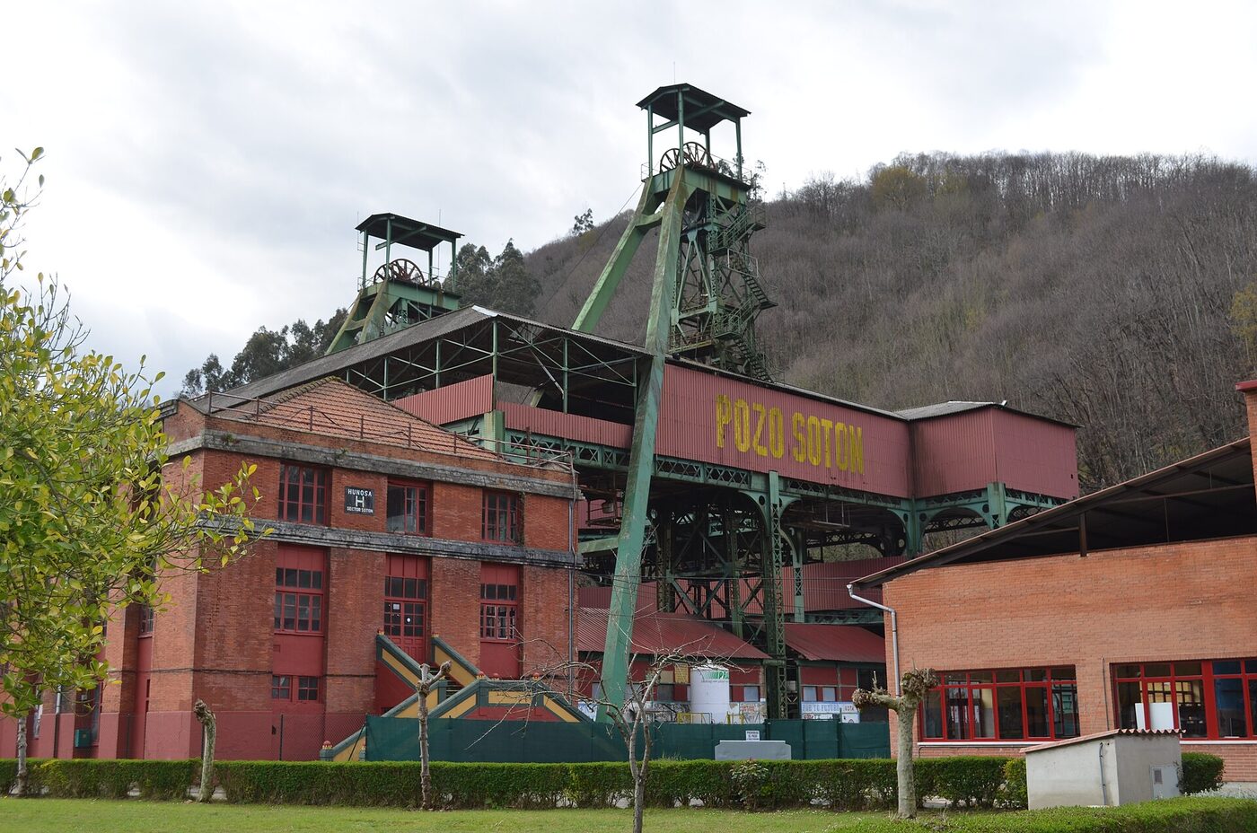 Two green steel winding headframes rising over a colliery, with red-painted and brick pithead buildings below lettered POZO SOTON in yellow, and a wooded hillside behind.