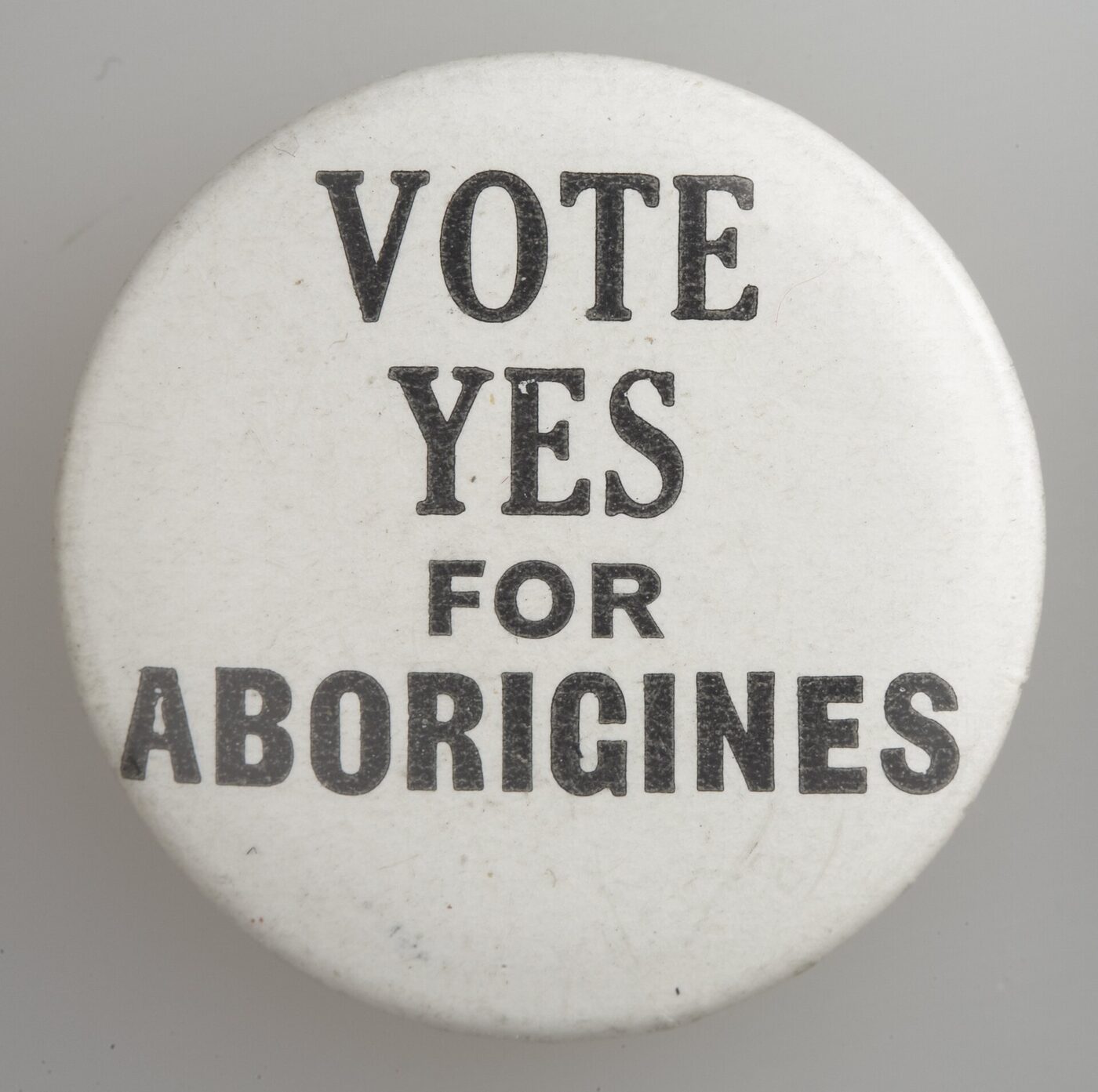A round white pin-back badge photographed against a plain grey ground, printed in black capitals with the words VOTE YES FOR ABORIGINES.